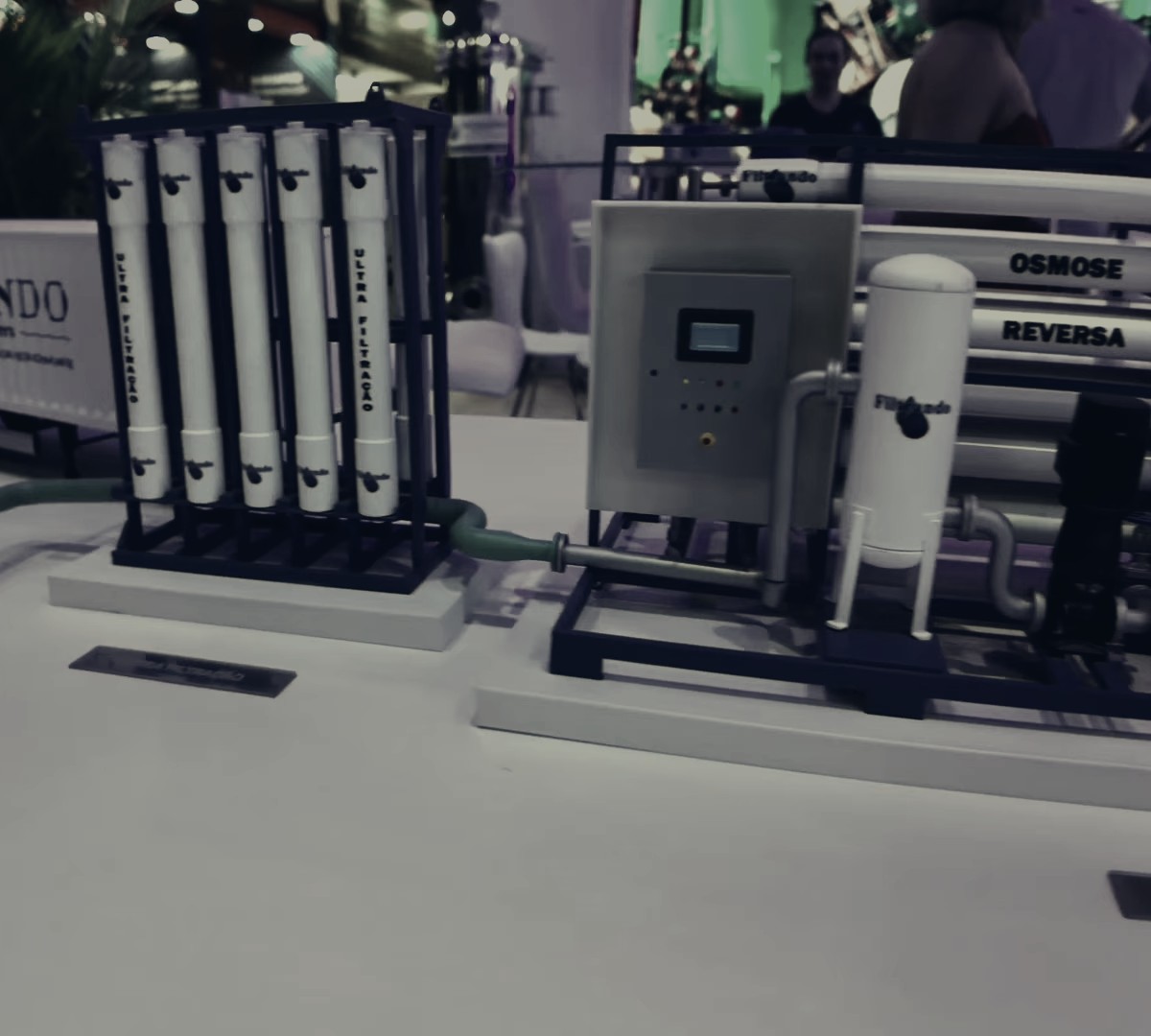Scale models of a water treatment plant on a booth table at Fenasucro: the ultrafiltration rack and the reverse osmosis skid, with the show lights behind.