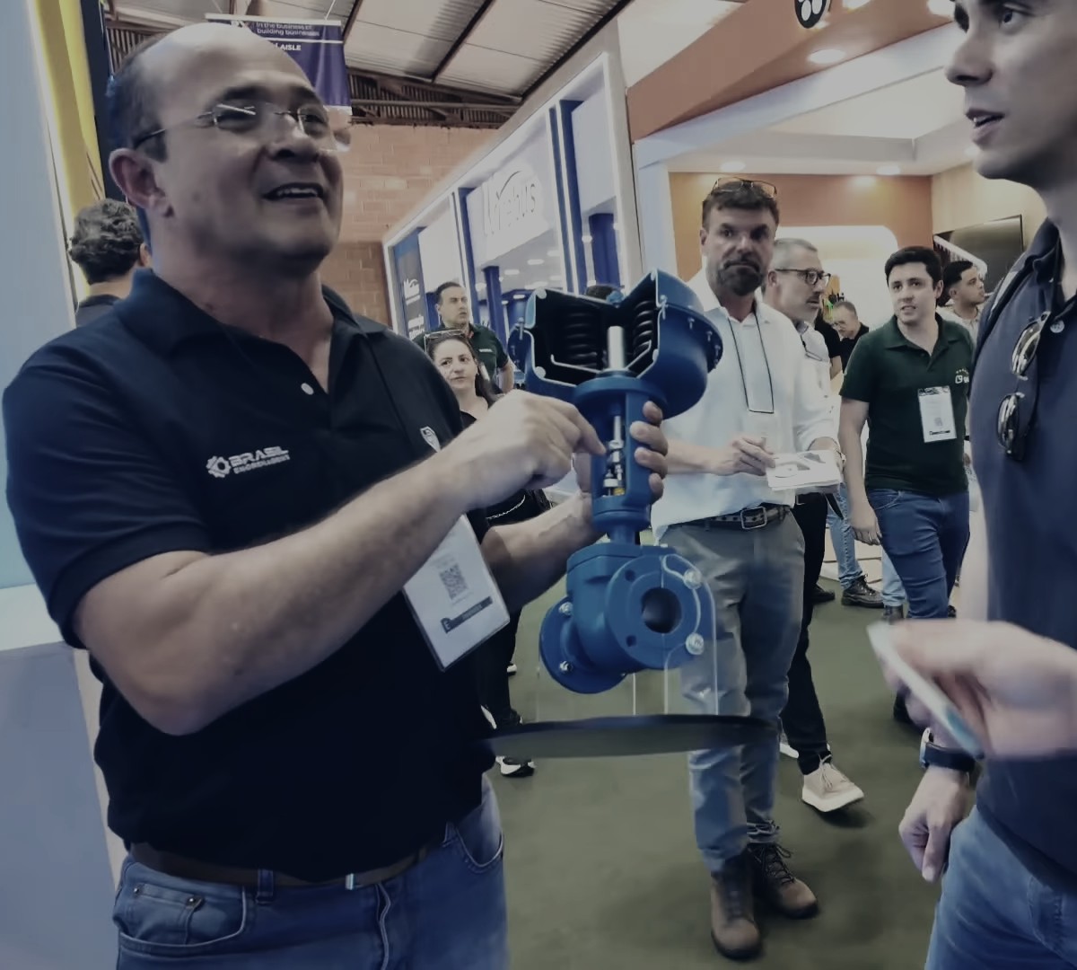An exhibitor holds a cutaway model of a control valve and shows the inside to a small group in the booth aisle.